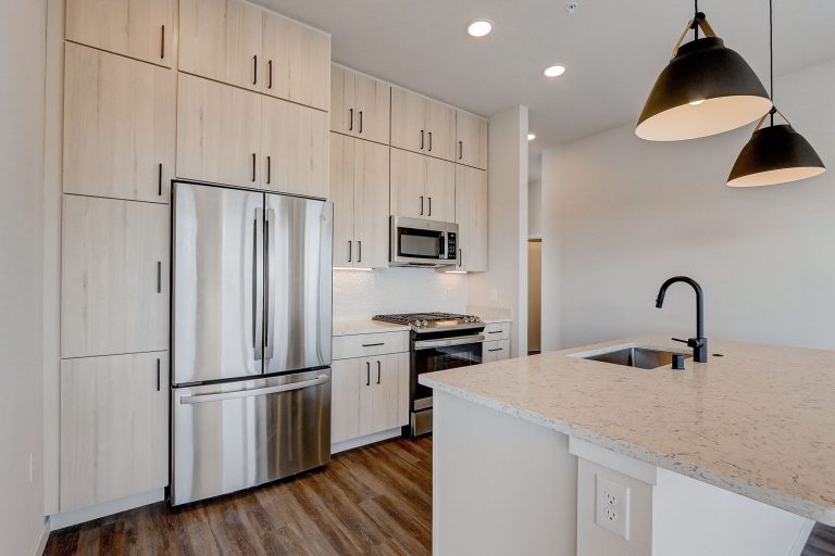 Modern kitchen with light wood cabinets, stainless steel appliances including a French door refrigerator, and a large island featuring a sink and black faucet. Pendant lights hang above the island