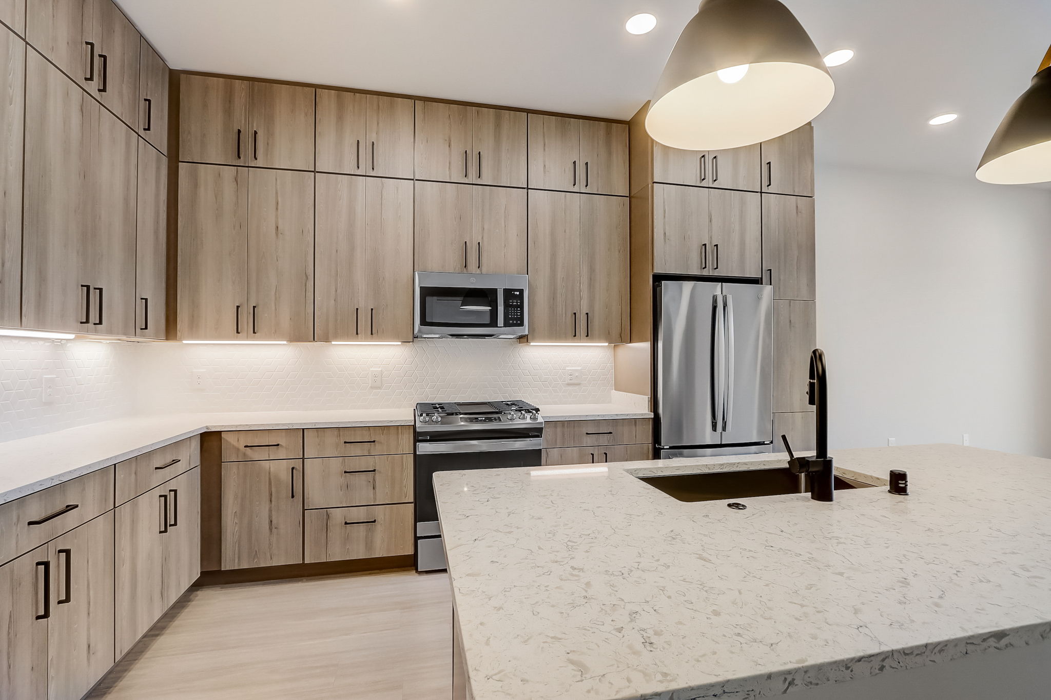 Modern kitchen with light wood cabinetry, stainless steel appliances including a microwave and refrigerator, and a large white marble island with an integrated sink. The space is well-lit with several recessed lights and pendant lights over the island.