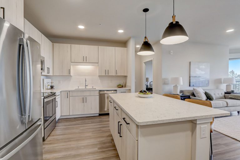 A modern kitchen with light wooden cabinets, stainless steel appliances, an island with a white countertop, and two pendant lights hanging above. The kitchen opens to a living area with light-toned furniture and large windows