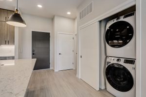 A modern laundry room features a stacked washer and dryer in a closet with sliding doors. The room has a light marble countertop, light wood flooring, and recessed lighting. A black pendant light hangs from the ceiling, and there are multiple doors leading out