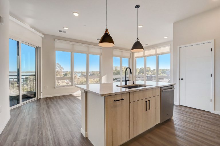 Modern kitchen and dining area with large windows offering ample natural light. The space features light wood cabinetry, a central island with a sink, stainless steel appliances, and a view of a balcony with sliding glass doors and wood flooring throughout