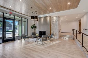 A modern, well-lit lobby area with a wooden ceiling and light wooden floors. The space features a seating area with four chairs around a glass coffee table, a large potted plant, and two black hanging light fixtures. Glass doors in the background lead outside