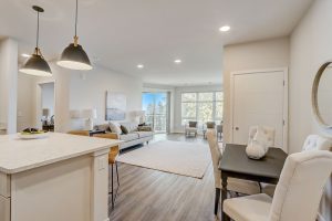 Modern open-plan living space with a kitchen island and pendant lights on the left, a dining area with a round mirror and seating for four on the right, and a cozy living area with a large window in the background. Light-colored walls and flooring create a bright atmosphere