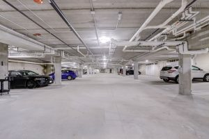 An empty underground parking garage with concrete pillars and pipes along the ceiling. Several cars are parked on the left and right sides of the spacious area, which is illuminated by overhead fluorescent lights