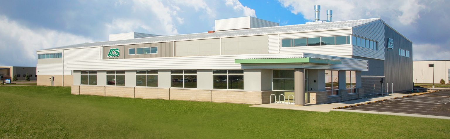 industrial building with a silver and white facade is set against a partly cloudy sky. The building features large windows and a green logo on the side.