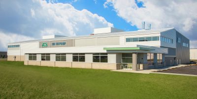 industrial building with a silver and white facade is set against a partly cloudy sky. The building features large windows and a green logo on the side.