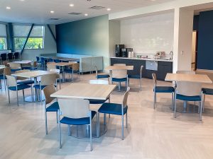 A bright, modern cafeteria with several wooden tables and blue chairs. A counter at the back has a coffee machine and condiments. Large windows and a pastel color scheme create an inviting atmosphere.