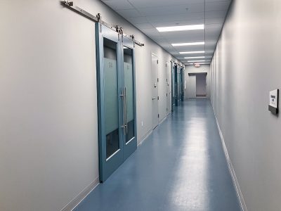 A long, empty hallway with blue flooring and ceiling lighting. On the left, there are teal sliding doors with glass panels