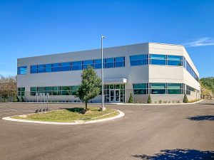 A modern, two-story office building with large windows and a flat roof under a clear blue sky. It's surrounded by a parking area, landscaping, and a single tree in the foreground.