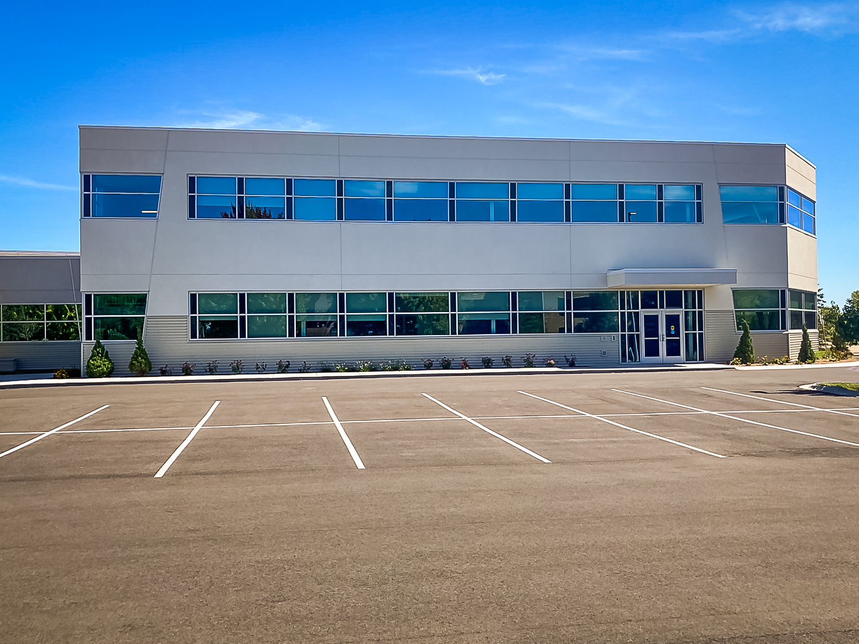 A modern, two-story office building with large windows and an empty parking lot in front. The sky is clear and blue, suggesting a sunny day.