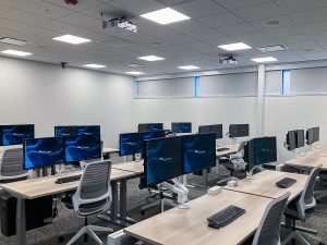 A modern, empty computer lab with multiple workstations. Each station has dual monitors, keyboards, and mice. The room features gray carpet, white walls, and a drop ceiling with fluorescent lights.