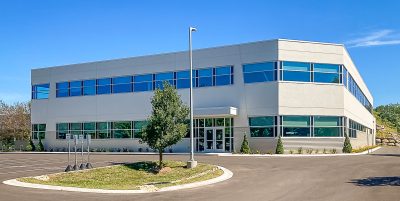 A modern, two-story office building with large windows and a flat roof under a clear blue sky. It's surrounded by a parking area, landscaping, and a single tree in the foreground.