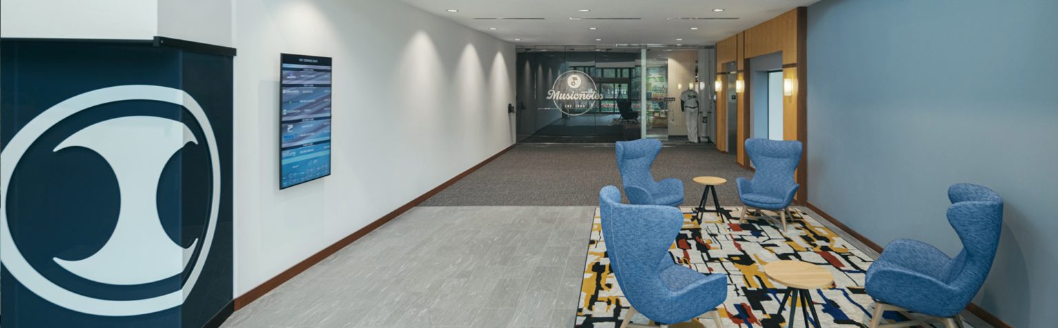 A spacious, modern office lobby with blue and white walls, featuring a central area with four blue armchairs around small wooden tables on a patterned rug. The rear wall displays a large logo.