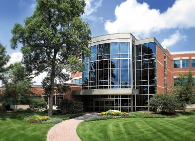 A modern office building with large glass windows and a red brick facade. Lush green grass and a variety of trees surround the structure, with a winding path leading to the entrance.