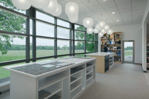 A well-lit room with large windows overlooking a grassy field. The room features minimalist white shelving units and high countertops. Several white spherical pendant lights hang from the ceiling.