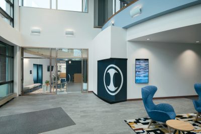 A modern office lobby with a gray floor, white walls, and large windows. The space features a seating area with blue chairs and a patterned rug.