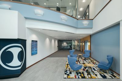 A spacious, modern office lobby with blue and white walls, featuring a central area with four blue armchairs around small wooden tables on a patterned rug. The rear wall displays a large logo.