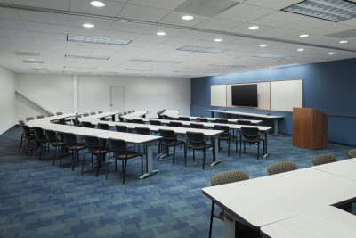 A large, empty conference room with blue carpet and white walls. It features a U-shaped arrangement of tables and chairs, a podium at the front right, and a large screen and whiteboard on the wall. Ceiling lights illuminate the room.