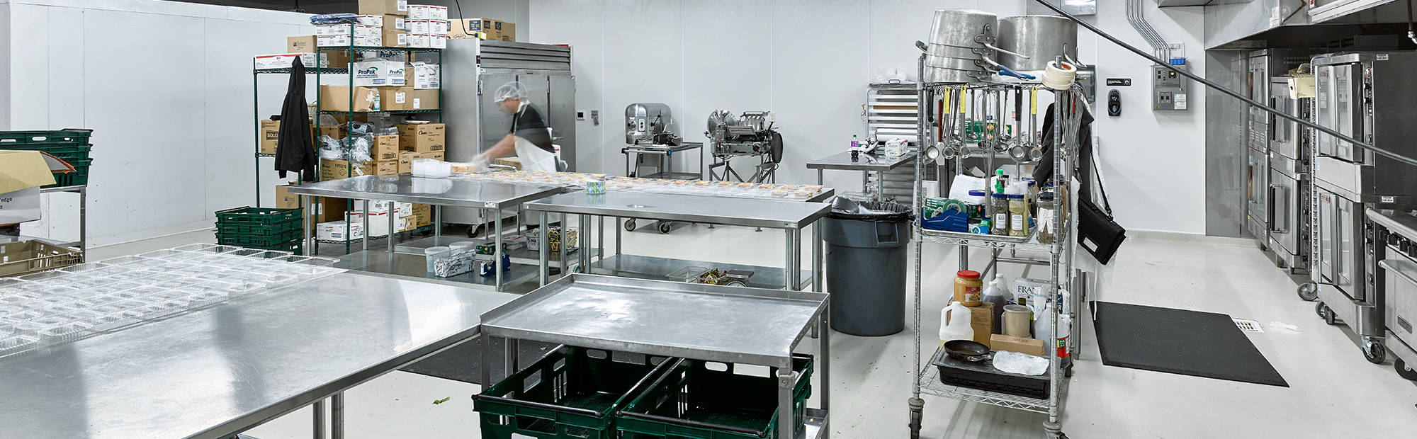 A commercial kitchen with stainless steel tables, shelves stocked with supplies, and industrial appliances. A person in the background is preparing food near trays and utensils. The area is clean and brightly lit.