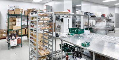 Industrial kitchen storage area with metal shelves and racks holding boxes, trays of pastries, and various supplies. Stainless steel tables are in the center, with a blurred motion figure suggesting movement. Bright overhead lighting illuminates the room.