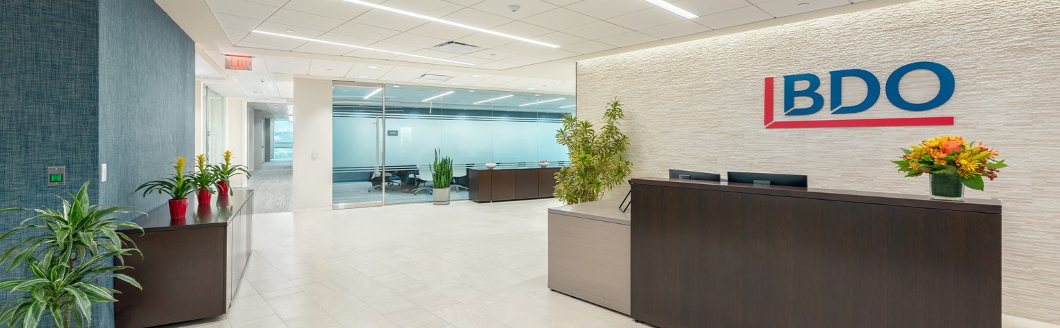 A modern office reception area features a large wooden desk with a black countertop, behind which is the BDO company logo. The space is decorated with potted plants, both on and near the desk. The floor has light-colored tiles, and a glass wall reveals a conference room.