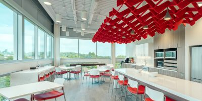 Modern office kitchen with white counters, red and white chairs, stainless steel appliances, and large windows overlooking greenery. Distinctive red geometric ceiling installation adds a vibrant touch. Ample seating includes bar stools and dining tables.