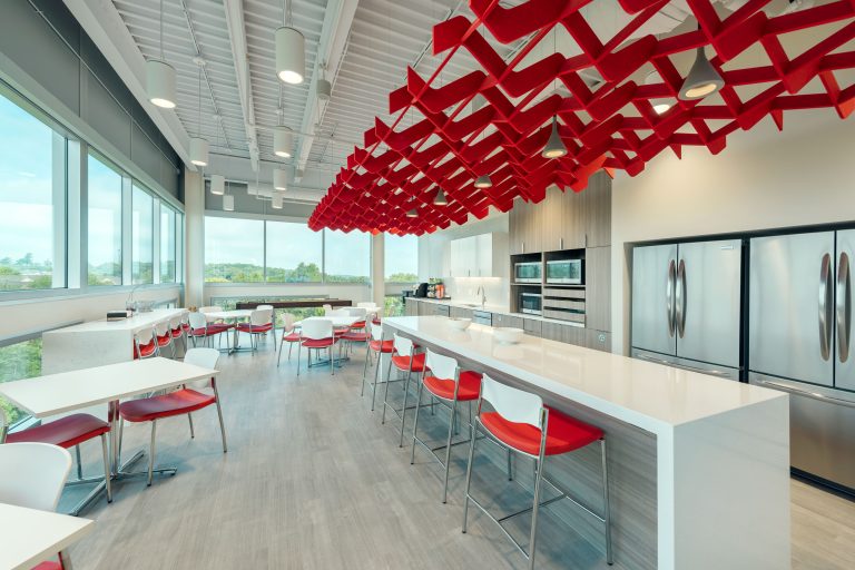 Modern office kitchen with white counters, red and white chairs, stainless steel appliances, and large windows overlooking greenery. Distinctive red geometric ceiling installation adds a vibrant touch. Ample seating includes bar stools and dining tables.