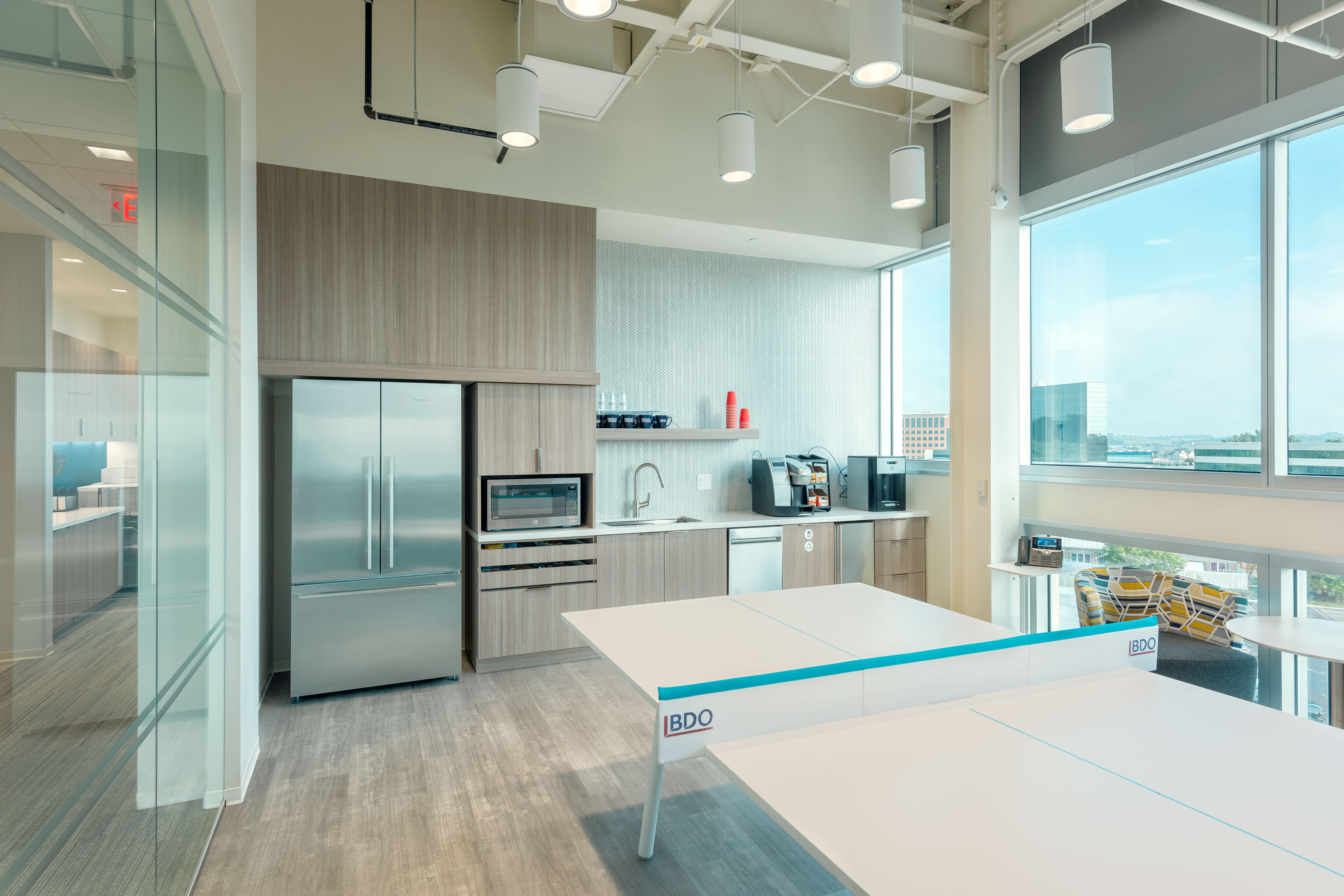 A modern office kitchen with wooden cabinetry, a refrigerator, microwave, sink, and coffee machine. Next to the kitchen is a ping pong table with "BDO" branding. The space is well-lit with large windows and overhead lights, and features city views outside.