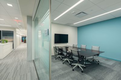 A modern, well-lit conference room with a large glass wall, featuring a dark wooden table and eight ergonomic chairs. A wall-mounted TV is at the head of the room. The adjacent hallway is visible, showcasing a clean, minimalist office design with white walls and carpeted floors.