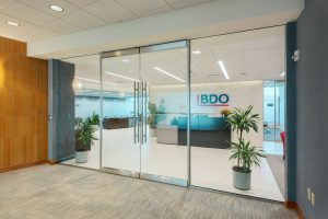 A modern office entrance with large glass doors, flanked by potted plants. Inside, there's a reception desk with the BDO logo on the wall behind it. The space is well-lit, featuring light-colored flooring and a mix of wooden and blue accent walls.