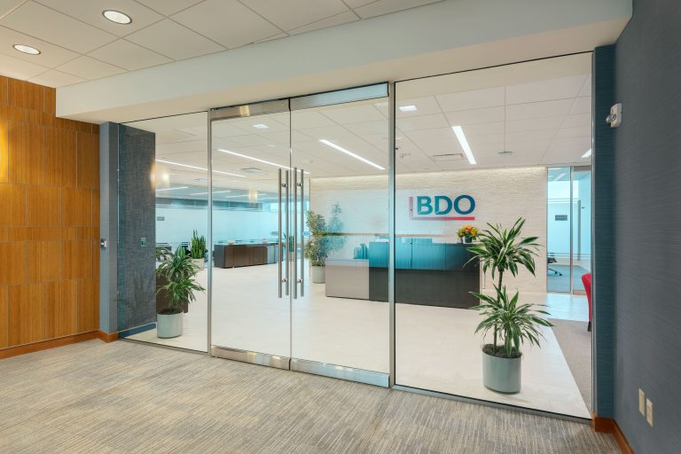 A modern office entrance with large glass doors, flanked by potted plants. Inside, there's a reception desk with the BDO logo on the wall behind it. The space is well-lit, featuring light-colored flooring and a mix of wooden and blue accent walls.