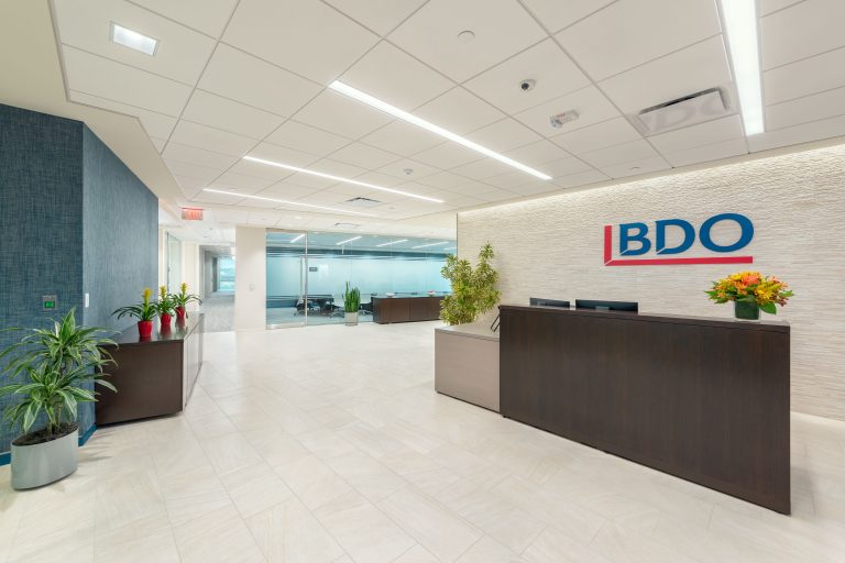 A modern office reception area features a large wooden desk with a black countertop, behind which is the BDO company logo. The space is decorated with potted plants, both on and near the desk. The floor has light-colored tiles, and a glass wall reveals a conference room.