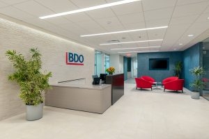 The modern reception area of an office with a BDO logo on the wall. The area features a reception desk, potted plants, and a seating area with two red chairs and a small table, positioned near a large window and a mounted TV screen on the wall.