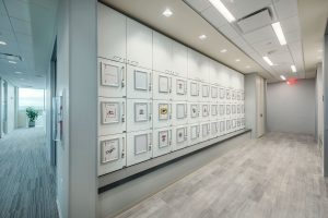 A modern office hallway featuring a wall of white lockers adorned with various logos and personal touches. The floor is made of light-colored wood, and the hallway leads to an office area with a large plant in the distance near a window. Ceiling lights are evenly spaced.