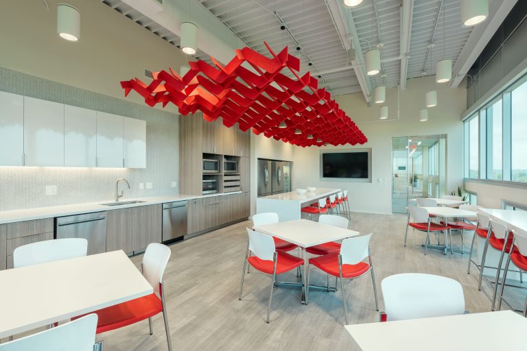 A modern office kitchen and dining area with white countertops, light wood cabinets, and stainless steel appliances. Red and white chairs surround white tables. A striking red abstract ceiling fixture contrasts with the light decor. Large windows bring in natural light.