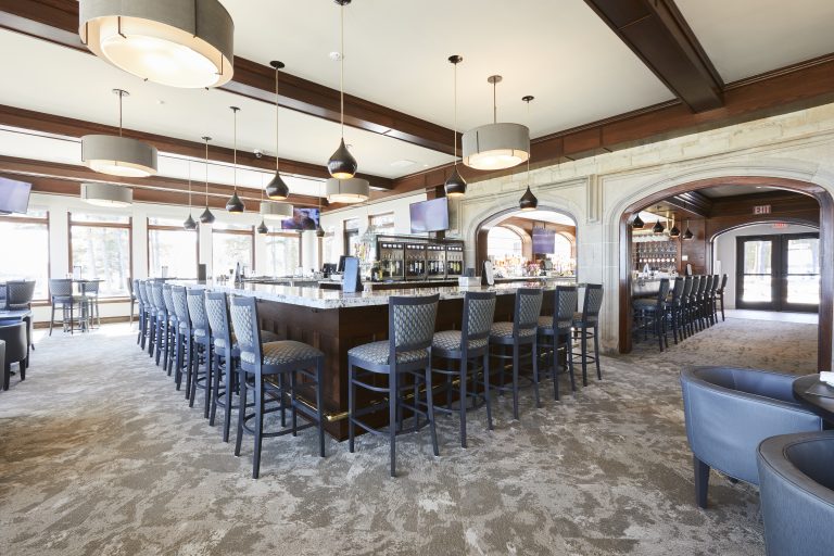 Upscale restaurant interior featuring a central rectangular bar with high-back chairs. Modern pendant lights hang from the ceiling. Large windows around the space allow natural light to pour in. Several seating areas are visible in the background.