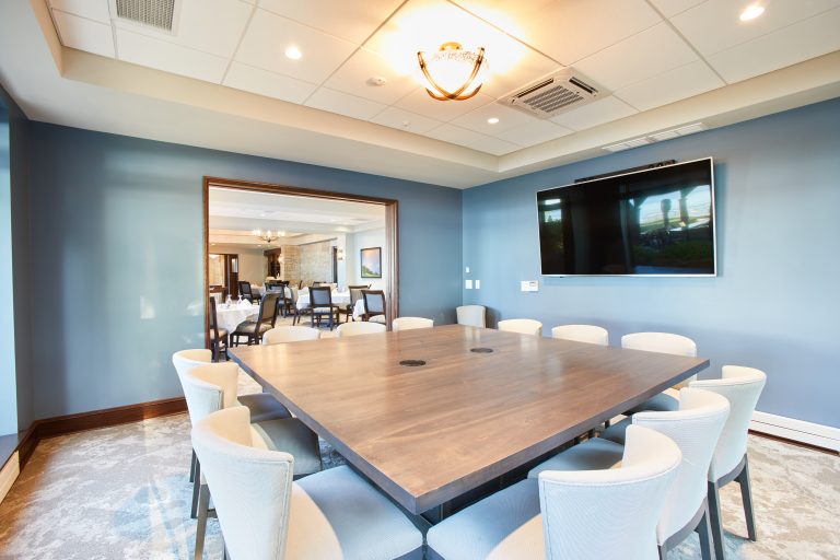 Country Club Conference room with square wood table, white fabric chairs, light blue walls, and a monitor on the wall