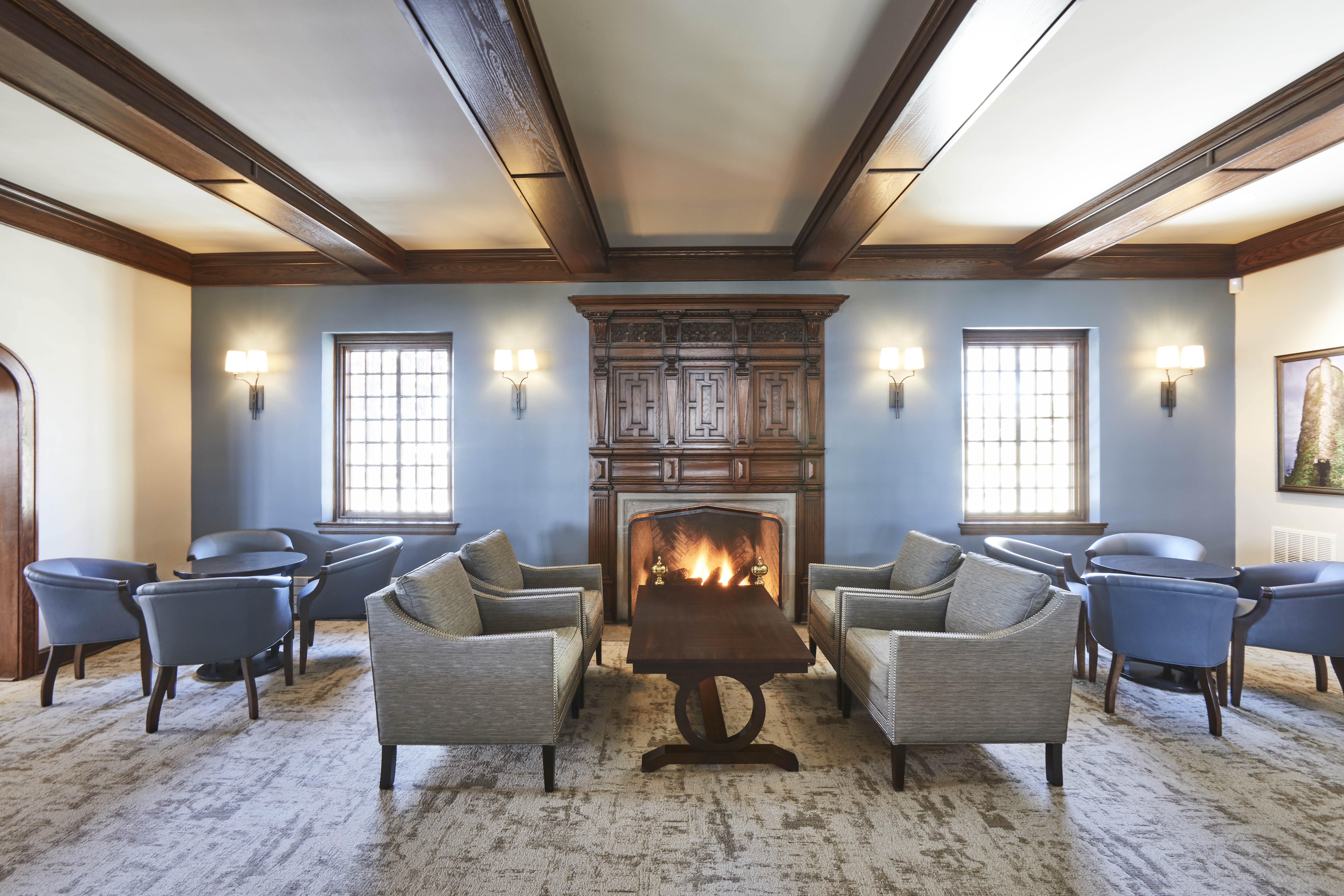 Lounge area with wood paneled fireplace in the center of the room, light gray fabric chairs, wall sconces, and carpeted floor with lattice windows