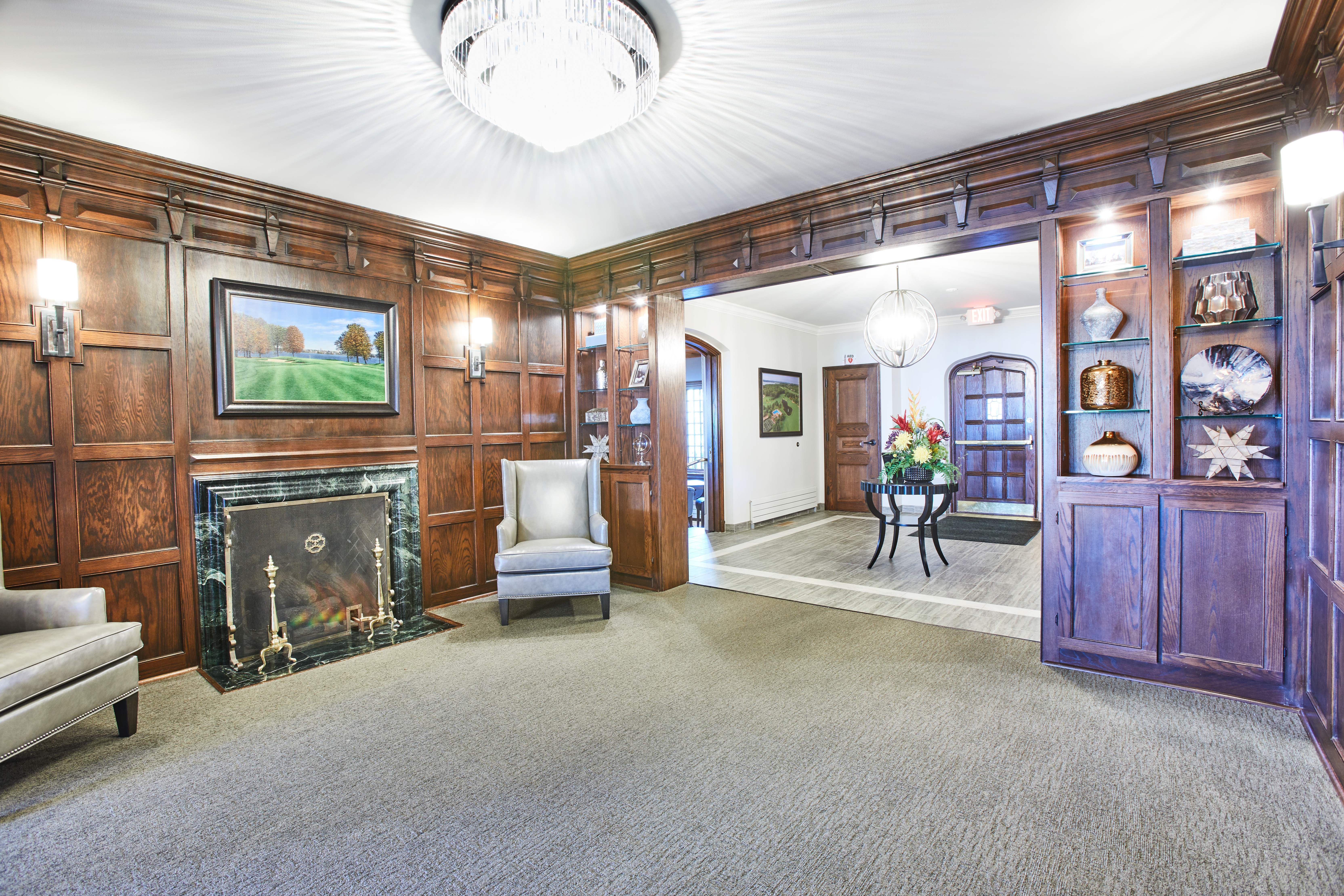 A luxurious room with wood-paneled walls, a fireplace, and a grand chandelier. The room has two gray armchairs positioned near the fireplace. Shelves inset into the walls display decorative items. A carpeted floor leads to an adjacent sunlit room through an arched doorway.