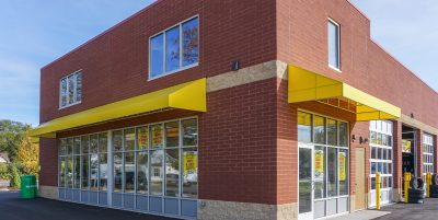 A brick industrial building with large glass windows and bright yellow accents, possibly a store or service center. Yellow caution lines are painted on the pavement leading to the entrance.