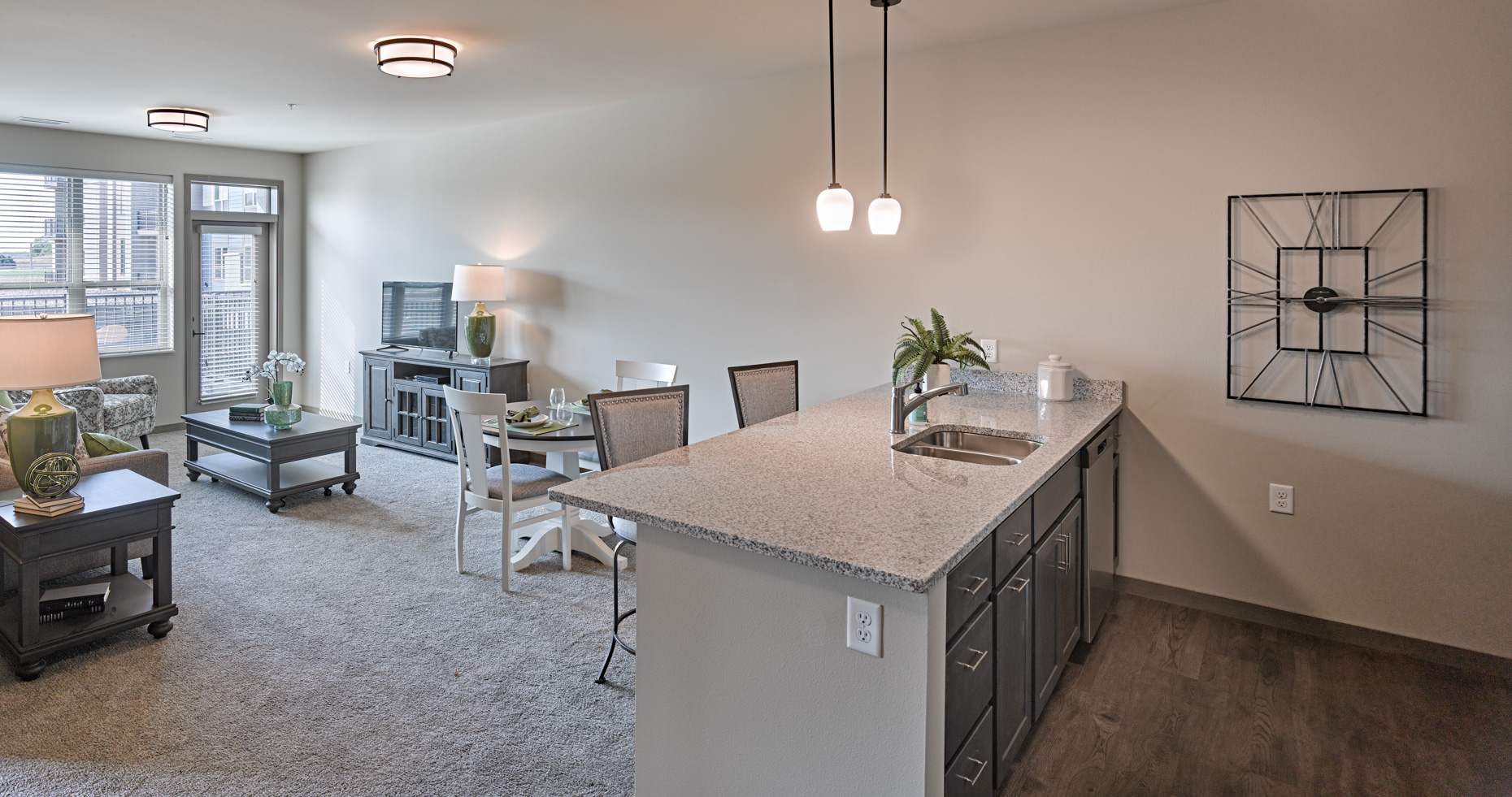 A modern, open-concept independent senior living apartment with a kitchen area. The living room has a gray couch, a coffee table, a TV, and two lamps. The kitchen features a granite island with a sink, pendant lights, and dark cabinets. A dining table with chairs is set near the living space.