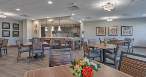 A well-lit, modern senior dining area features several square tables with striped chairs. Each table has a floral centerpiece. In the background, a kitchen area with pendant lighting, gray cabinets, and a sign that reads "The Kitchen" is visible. Artwork adorns the walls.