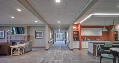 A cozy, modern communal area with a seating arrangement including armchairs, a small table, and a TV mounted above a fireplace. The hallway leads to a brightly lit room with a wall of cabinets on the right. The space features neutral tones and contemporary decor.