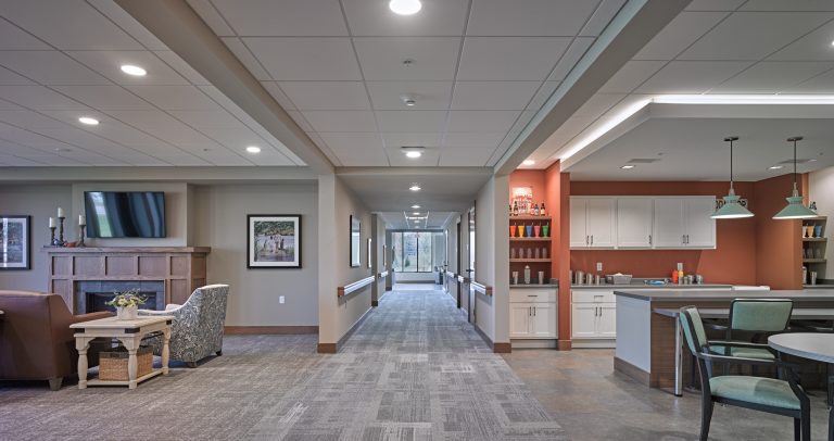 A cozy, modern communal area with a seating arrangement including armchairs, a small table, and a TV mounted above a fireplace. The hallway leads to a brightly lit room with a wall of cabinets on the right. The space features neutral tones and contemporary decor.