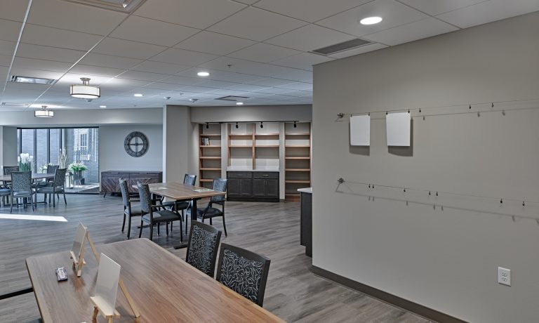 A modern, indoor community room with wooden floors and neutral-toned walls. The room features several tables and chairs, shelving units, and a bulletin board on the wall. Large windows on the left side allow natural light to enter the space.