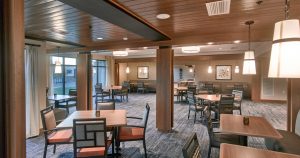 A modern dining area with wooden tables and chairs placed on a carpeted floor. Pendant and recessed lighting illuminate the space, which features warm wooden accents and large windows. Framed artwork decorates the walls, creating a cozy and inviting atmosphere.