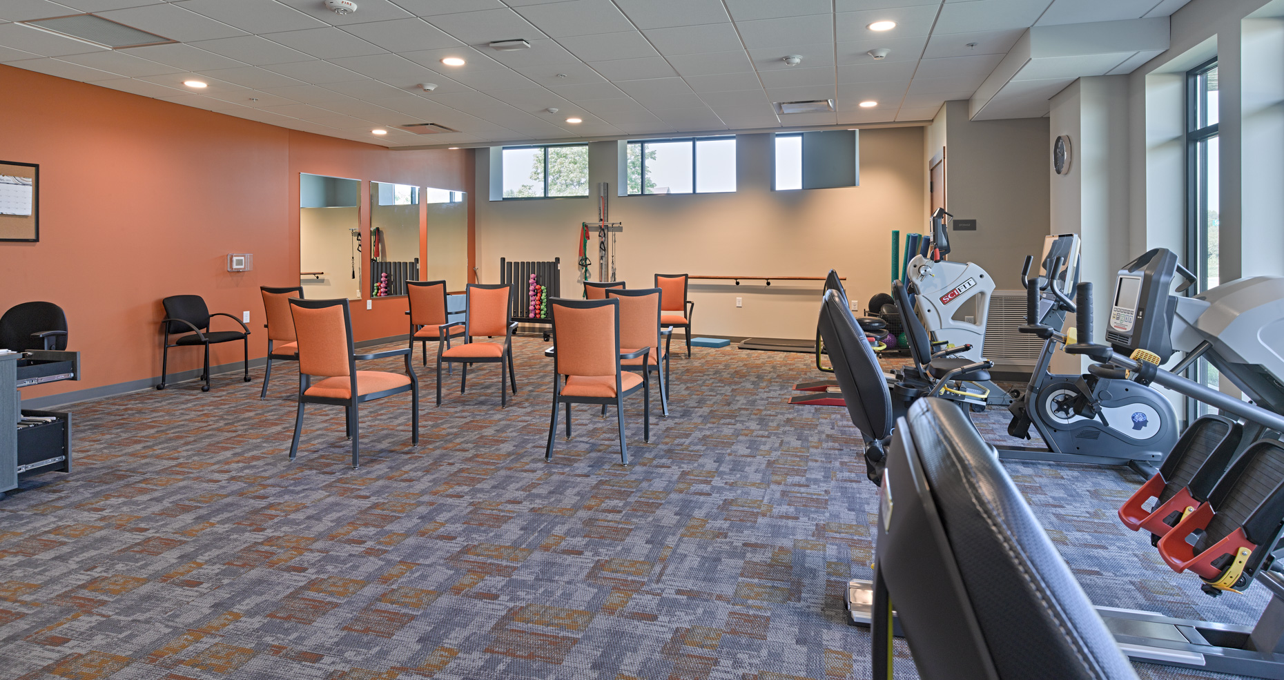 A well-lit fitness room with large windows features several exercise bikes, treadmills, and other gym equipment against the walls. Four orange chairs are arranged in the center of the room facing each other. The floor is carpeted with a multicolored pattern.