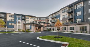 A modern multi-story senior living facility with balconies and large windows. The facade features a mix of grey, beige, and blue tones. There's a circular driveway and a few small trees in the foreground on well-maintained landscaping.