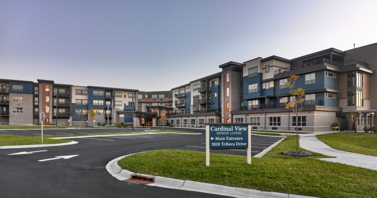 A modern, multi-story senior living facility with a sign in the foreground reading "Cardinal View Senior Living Main Entrance 3890 Tribeca Drive." The building has a combination of brick and siding with large windows and landscaped surroundings.