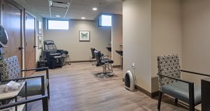 An interior view of a modern salon room with two styling chairs, a reclining chair, and two patterned armchairs. The room features wood flooring, a light-colored ceiling, and walls mounted with a framed butterfly artwork. Large windows allow natural light to enter.